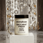 Luxury product photography of Aromahoes Baccarat Fresh Body Crème on a white marble plinth, surrounded by crystal prisms, a silver glitter telephone, and a sparkling 'Ah' jewelry charm.